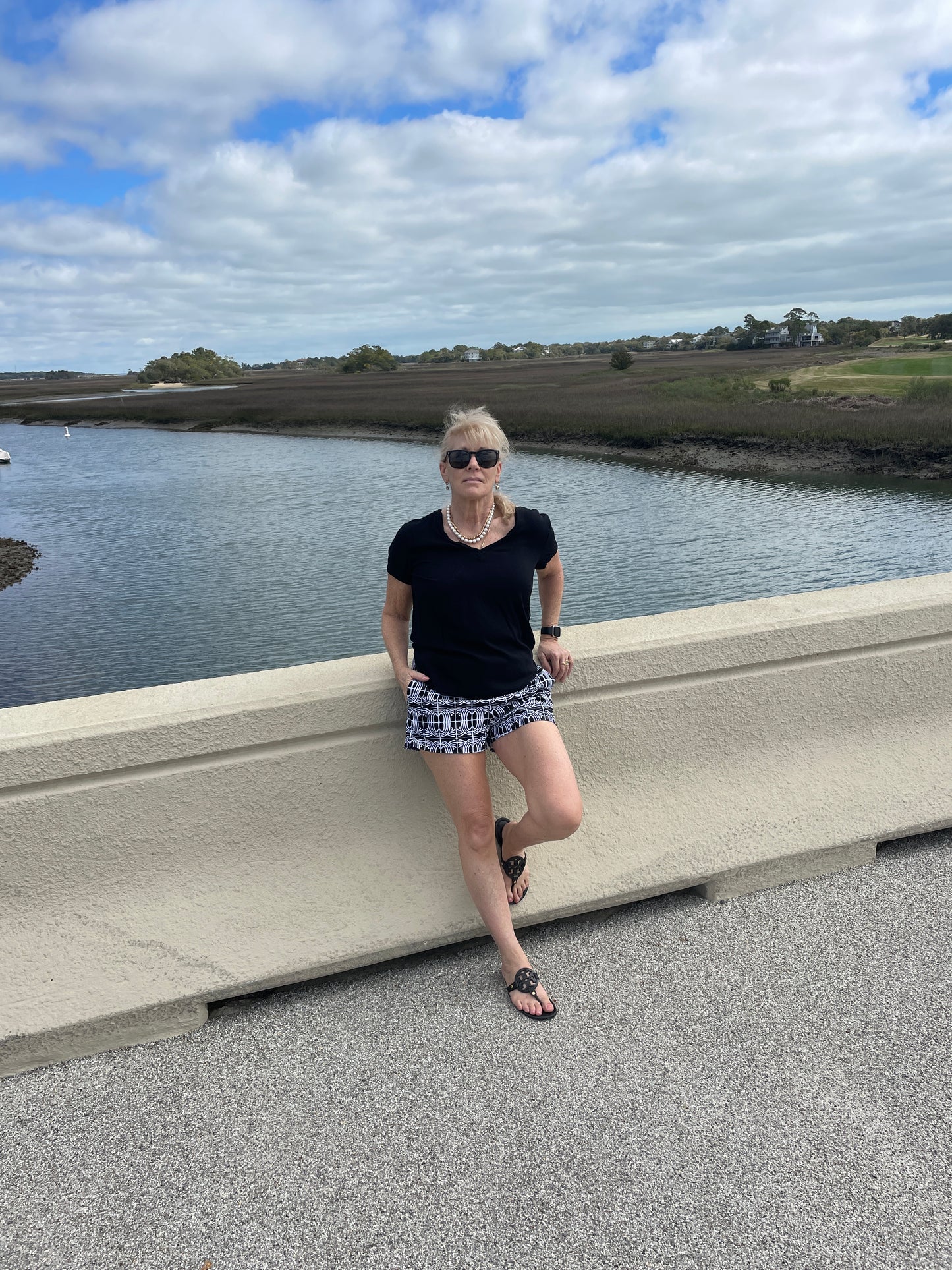 Woman resting on bridge with water in background wearing black and white shorts and solid black top with hand in pocket modeling product shorts